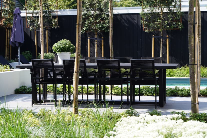 Zwarte eettafel met stoelen onder pergola, witte bloeiende bodembedekkers en zwembad