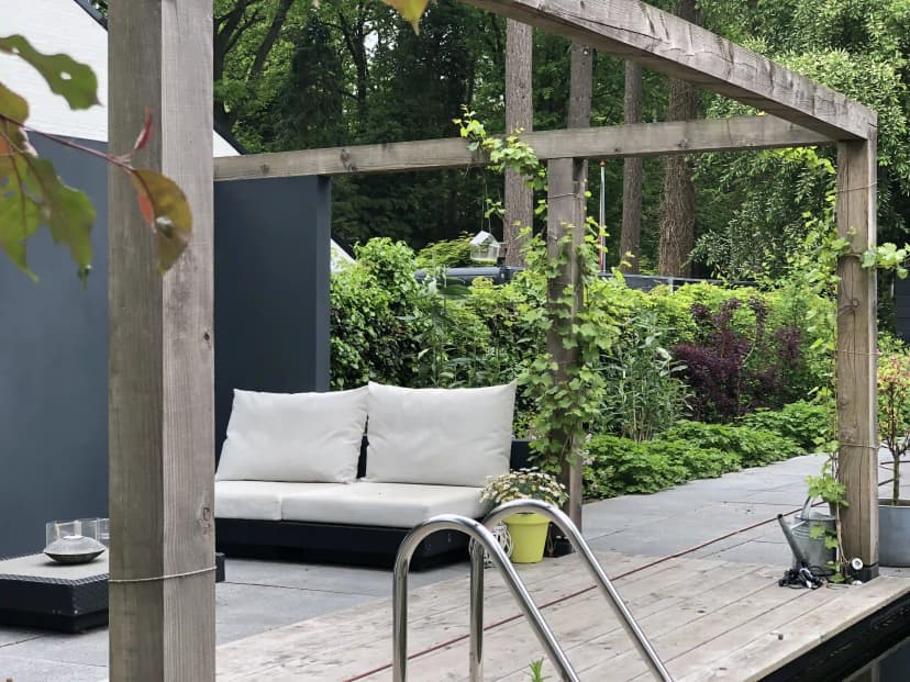 Overdekte zithoek met witte kussens onder houten pergola bij zwemvijver