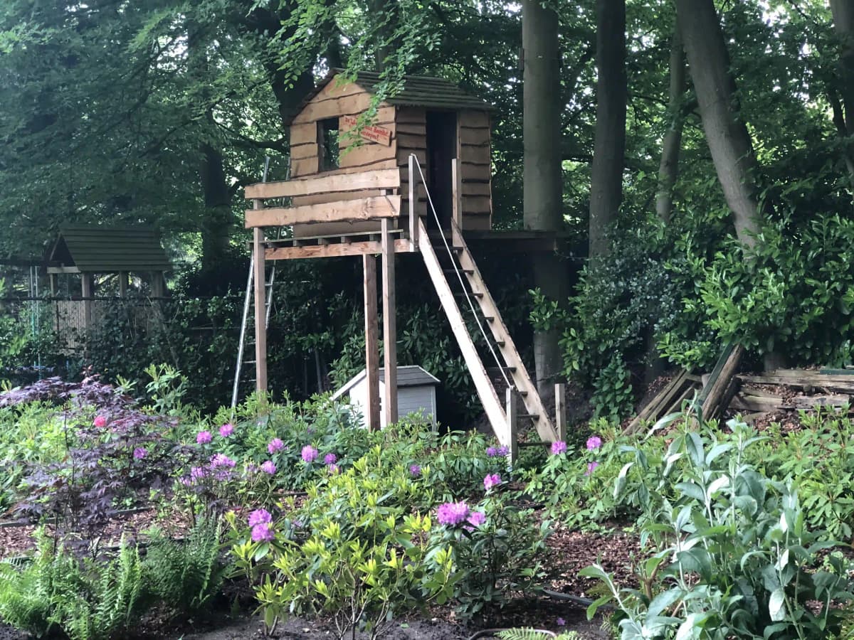 Houten speeltoestel met glijbaan omringd door paarse bloeiende planten en hosta's