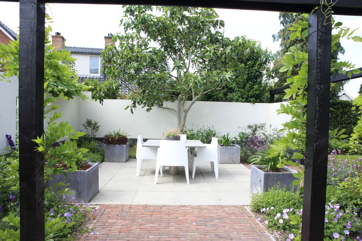 Omsloten binnentuin met witte eettafel onder donkere pergola en rijke beplanting